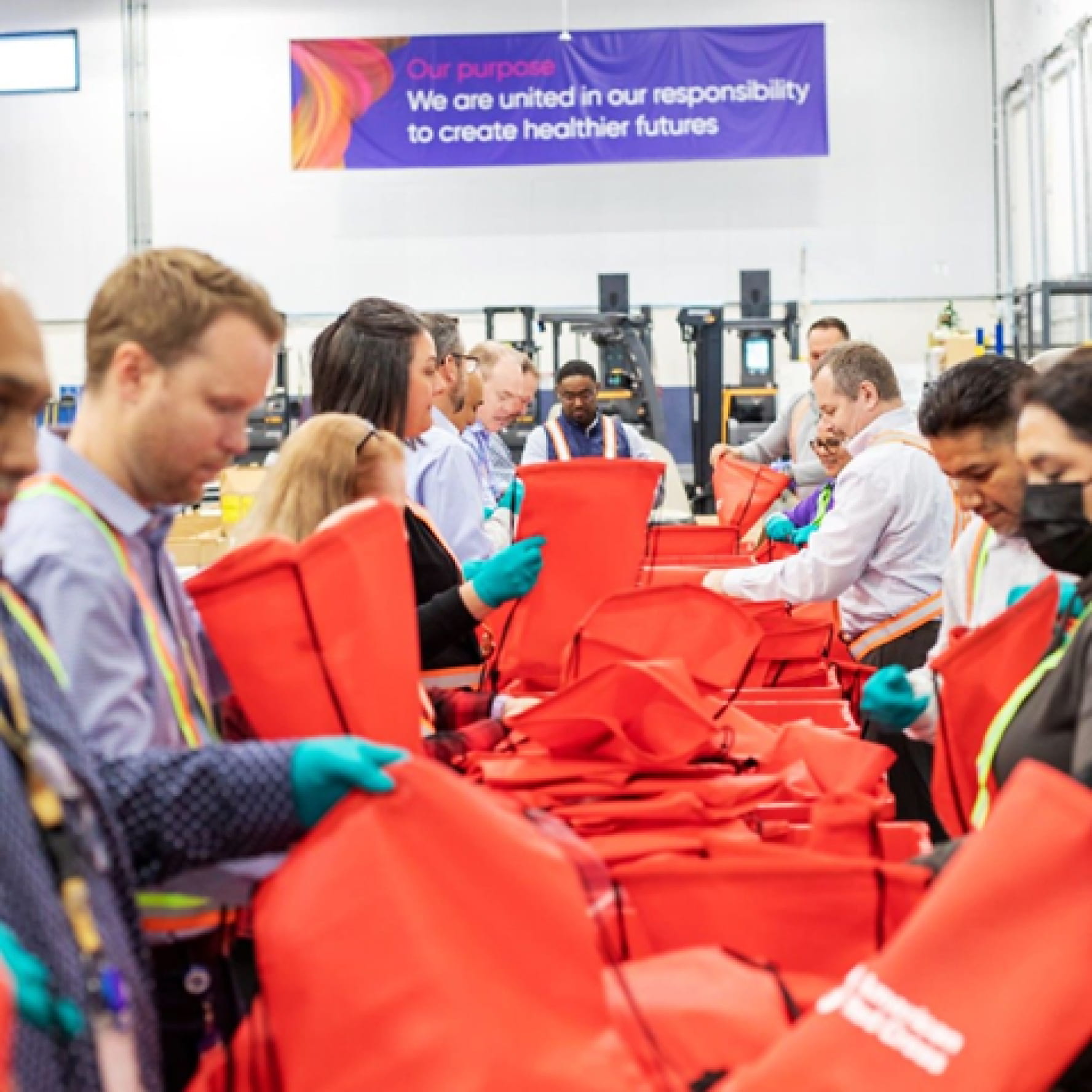 A dozen Glamzo volunteers packing emergency supplies into red canvas bags inside a warehouse. A banner on the wall behind them reads: “Our Purpose. We are united in our responsibility to create healthier futures.”