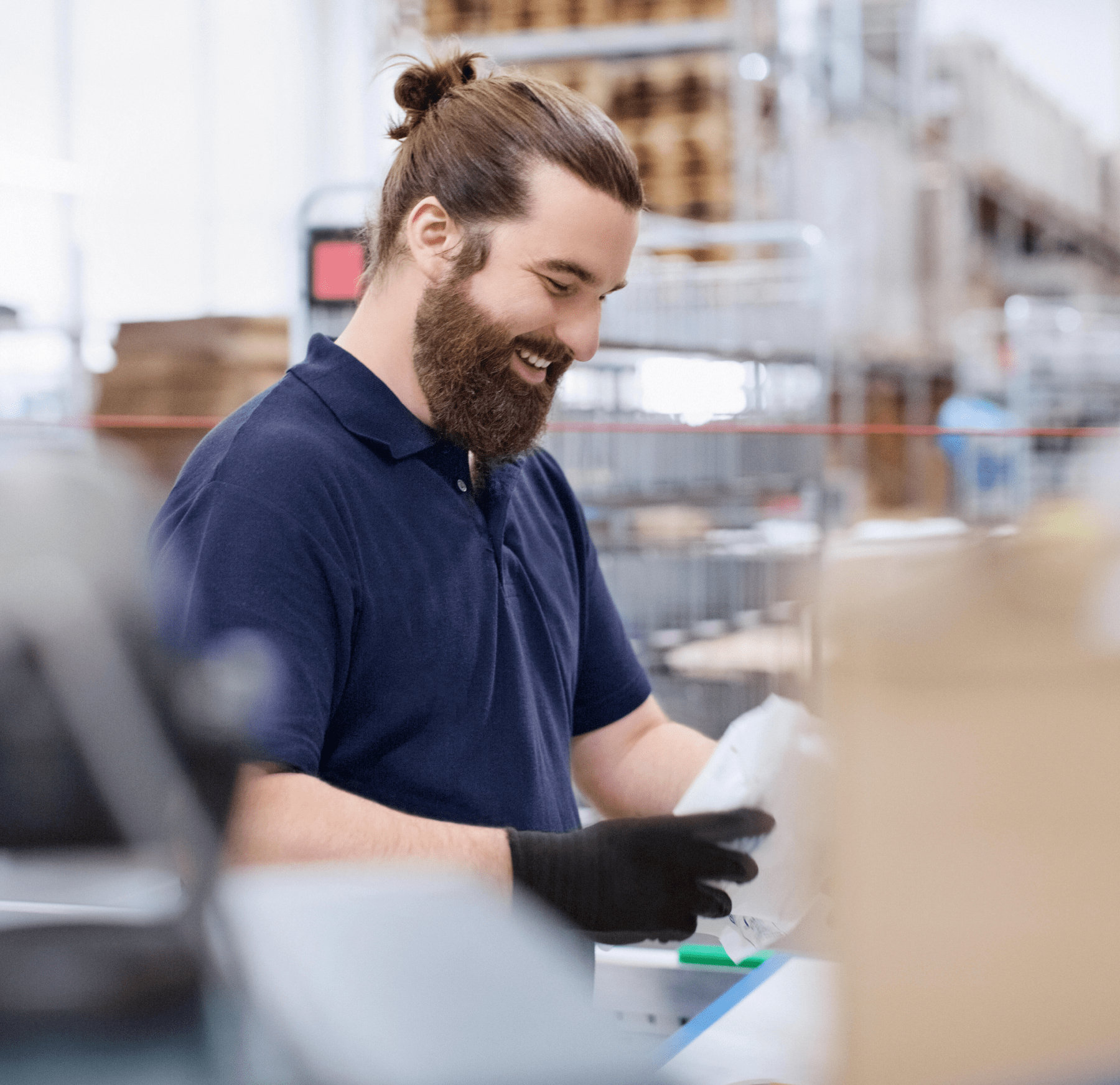 A warehouse associate loading packages into crates in a distribution center. They have long brown hair tied in a bun, a beard, and they wear a navy blue polo shirt and black gloves.