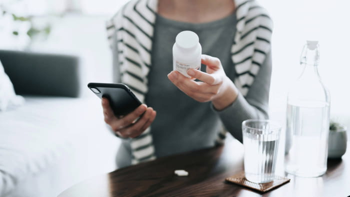 A person holding a white prescription medication bottle in one hand and a cell phone in another. They sit at a table with a glass of water in front of them while wearing a gray long-sleeve shirt and a black and white striped sweater.