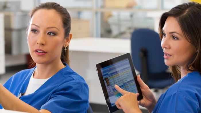 Two nurses sitting at a hospital desk discuss something while one holds and points at a tablet device. They both wear blue scrubs, have long dark hair, and medium skin.