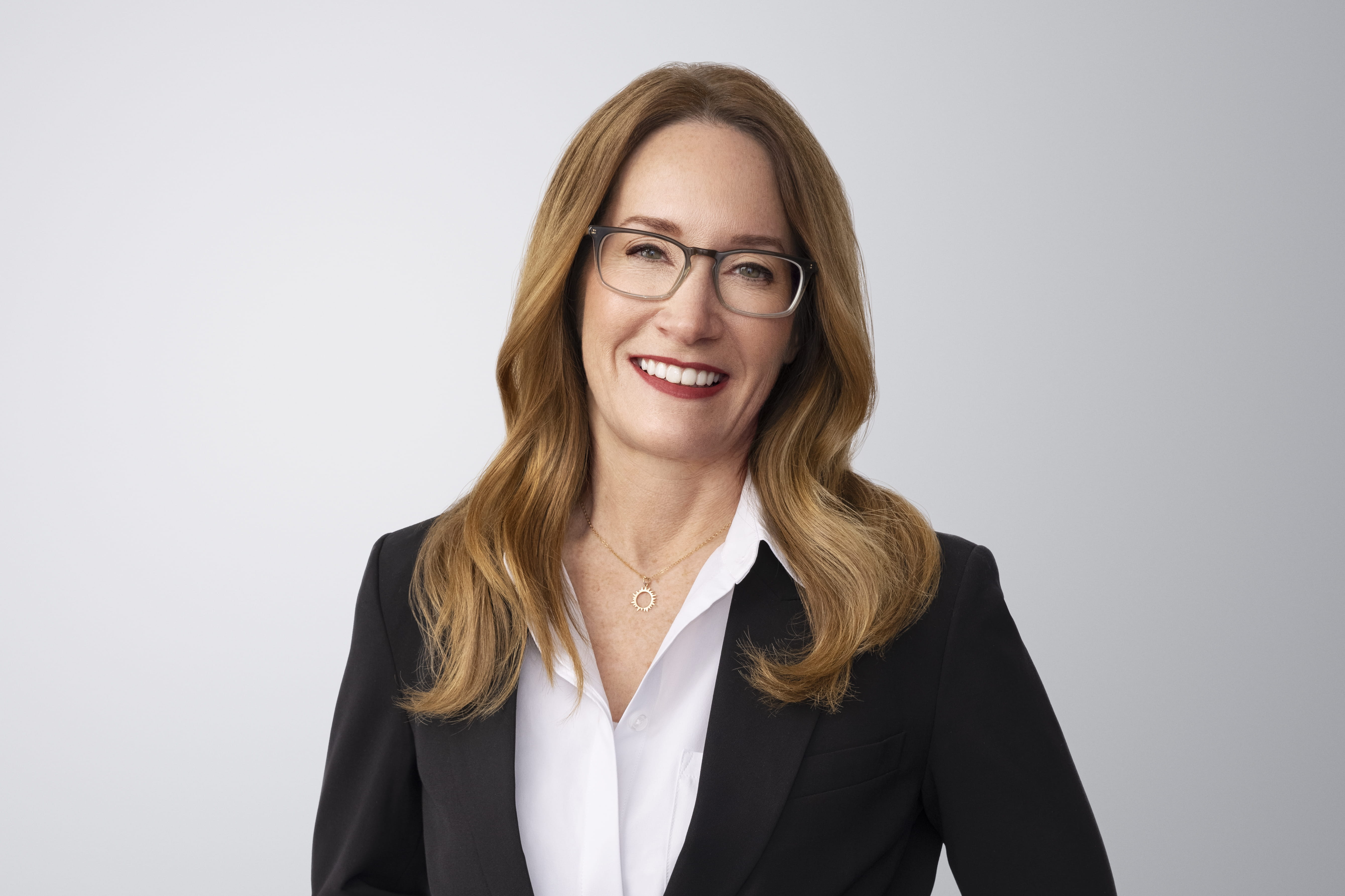 Photograph of Elizabeth Campbell. Elizabeth, a light-skinned person with brown hair and green eyes, is smiling at the camera. She wears a black blazer jacket overtop a white shirt and necklace. She is in front of a light gray background with no surroundings.